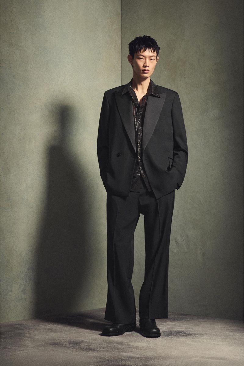 An Asian male stands in a full shot, dressed in a black suit and printed shirt, against a mottled green background.