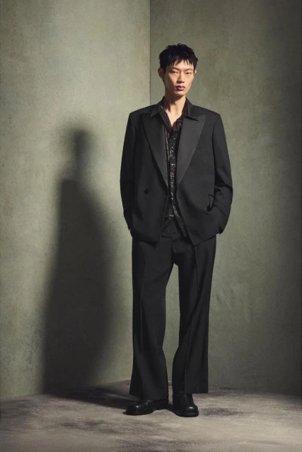 An Asian male stands in a full shot, dressed in a black suit and printed shirt, against a mottled green background.