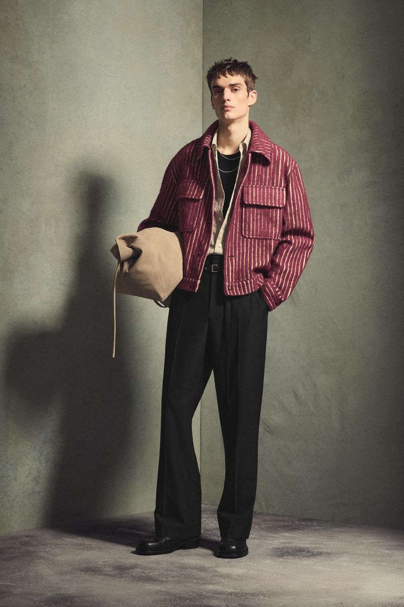 Full-length portrait of a male model wearing a red striped jacket and black pants while holding a beige bag in a studio setting.