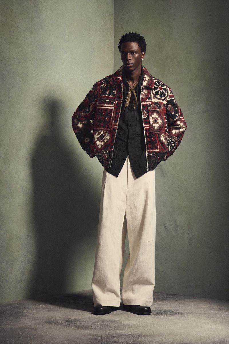 Man poses in patterned jacket and wide-leg pants in a studio setting, showcasing a modern fashion editorial shot with artistic styling.