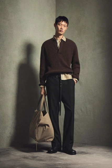 Studio shot of an Asian male model wearing a brown sweater and black dress pants while holding a beige tote bag against a textured wall.