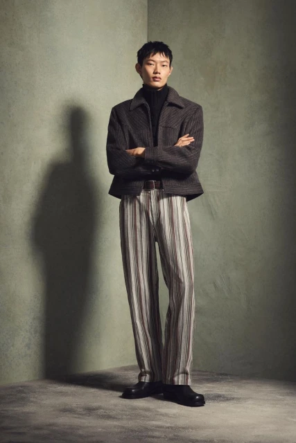 Full-length studio shot of a young adult male in a wool jacket, black turtleneck, and striped trousers, posing against a textured grey wall.