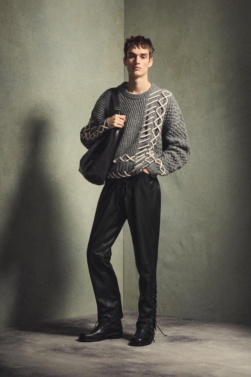 Full-length studio shot of a male model wearing a grey knit sweater, black leather-look pants, and black boots, carrying a shoulder bag.
