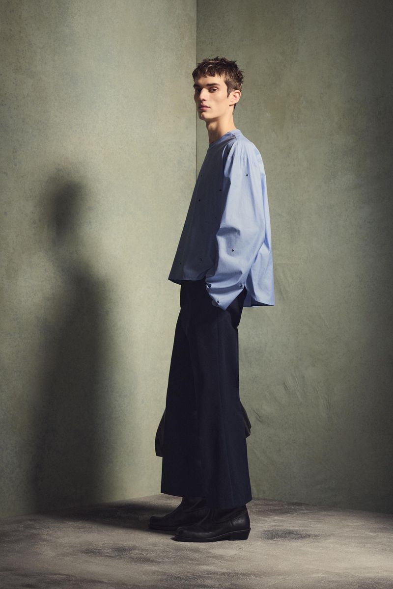 Fashion shot: a slender adult male model in an oversized light blue shirt and wide-leg navy pants poses indoors against a textured gray corner.