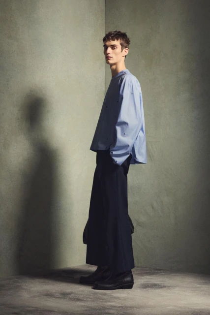 Fashion shot: a slender adult male model in an oversized light blue shirt and wide-leg navy pants poses indoors against a textured gray corner.