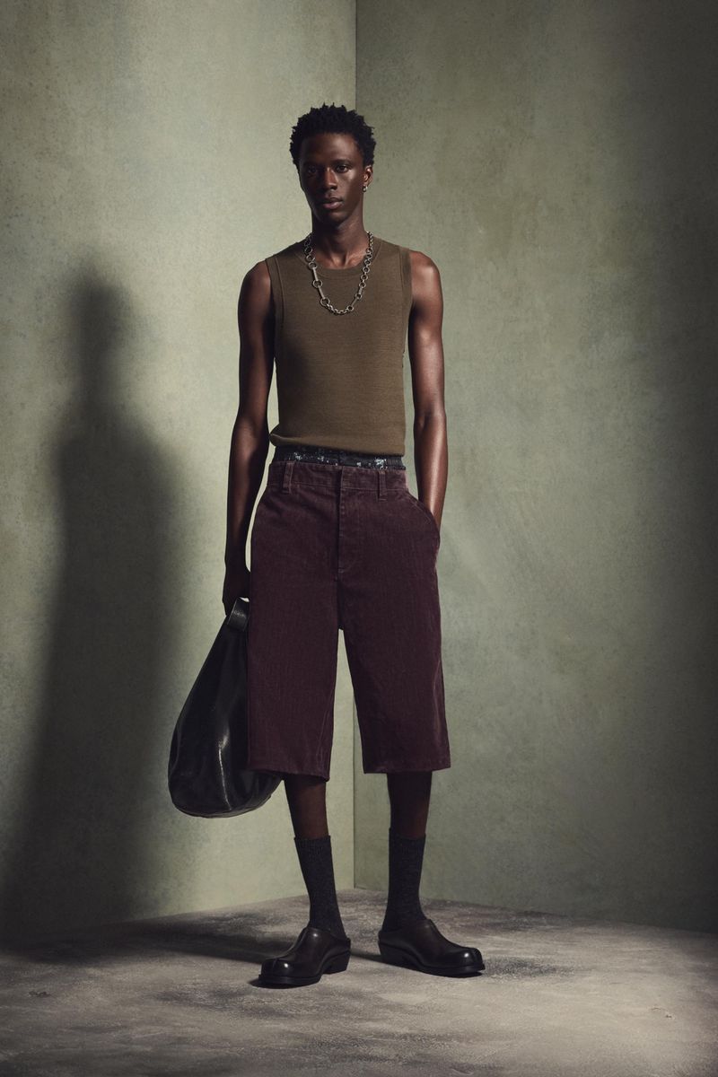A single dark-skinned adult male wearing a tank top and shorts stands indoors with a bag in hand against a textured greenish wall.