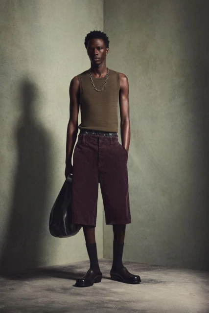 A single dark-skinned adult male wearing a tank top and shorts stands indoors with a bag in hand against a textured greenish wall.