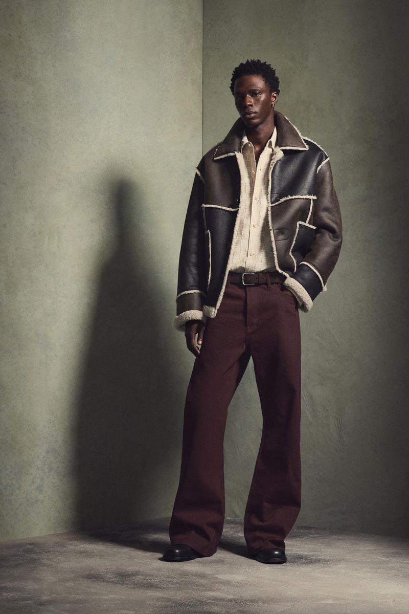 A Black male model in a shearling jacket, shirt, and wide-leg pants poses in a photo studio. The background is a muted green.