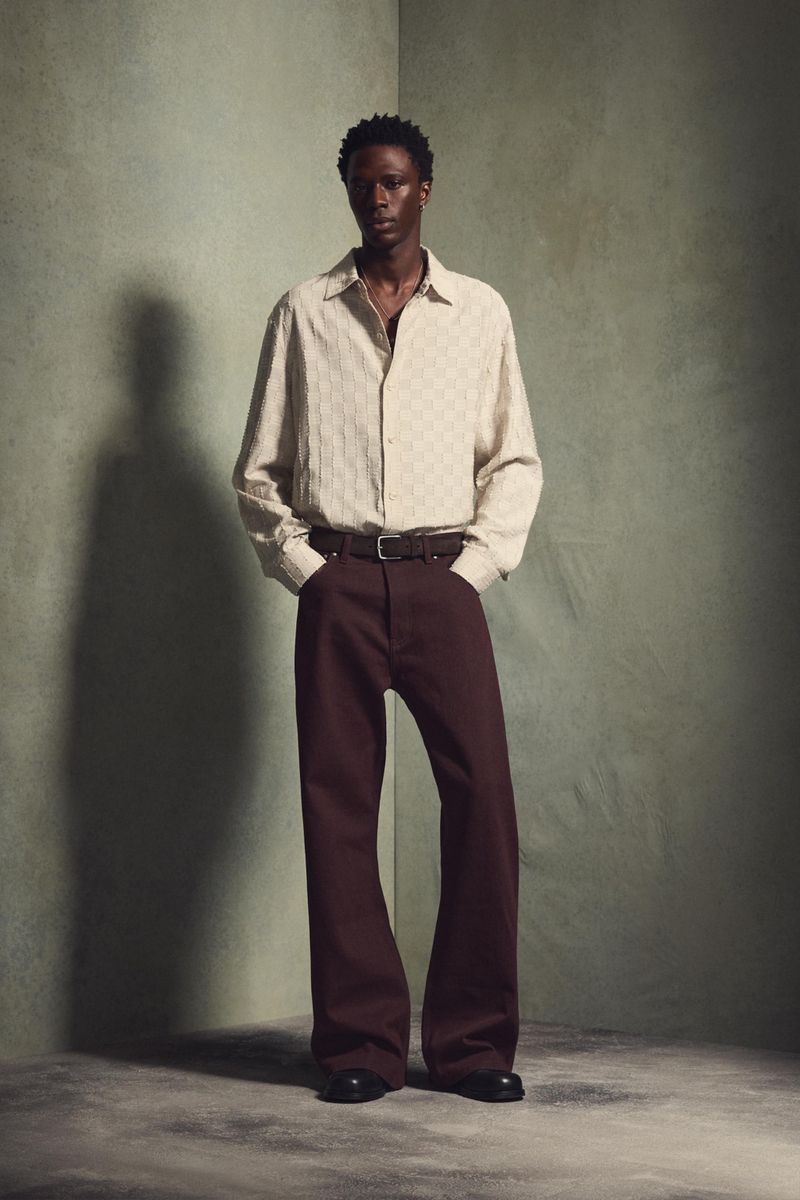Model wearing a textured cream shirt and brown trousers posing against a muted green studio corner background.
