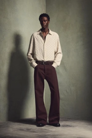 Model wearing a textured cream shirt and brown trousers posing against a muted green studio corner background.