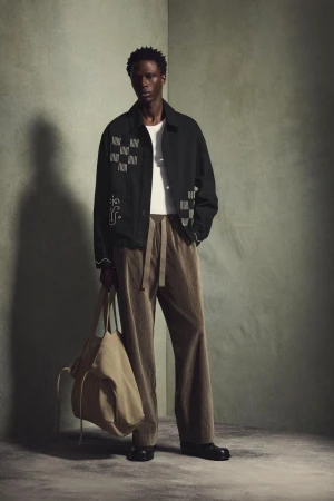 Model wearing a black jacket and brown trousers holding a large tote bag against a textured grey wall