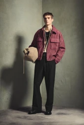 Full-length portrait of a male model wearing a patterned red jacket and black trousers holding a beige bag against a textured wall.