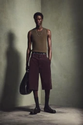 Model wearing an olive tank top and burgundy shorts holding a black bag against a muted wall.