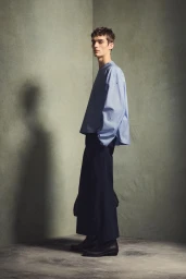 Full-length portrait of a male model wearing a light blue buttoned shirt and dark trousers against a textured grey studio wall.