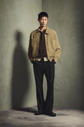Model in a tan suede jacket, dark brown knit polo, and black trousers standing against a textured studio wall.
