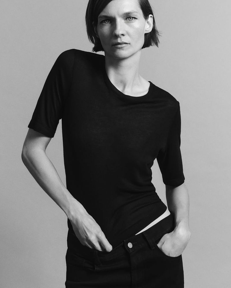 Model wearing a black t-shirt and dark trousers posing against a grey seamless studio background.