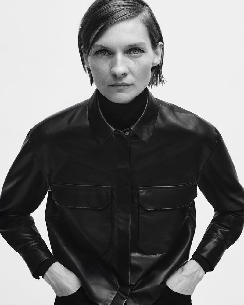A monochromatic portrait of a confident, fair-skinned woman with short hair, wearing a black leather shirt and turtleneck sweater.