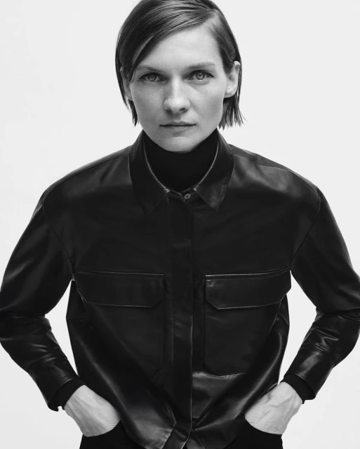 A monochromatic portrait of a confident, fair-skinned woman with short hair, wearing a black leather shirt and turtleneck sweater.