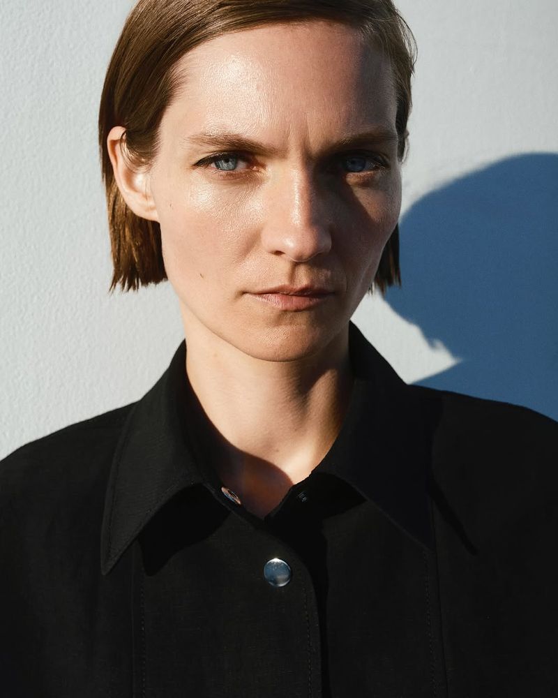 Headshot of an adult female with short brunette hair, wearing a black collared shirt in front of a light gray background with a shadow.