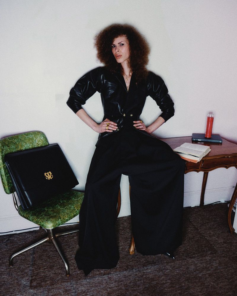 Stylish woman with afro sits on desk, wearing a black leather jacket, black pants, posing in an indoor setting with neutral background.