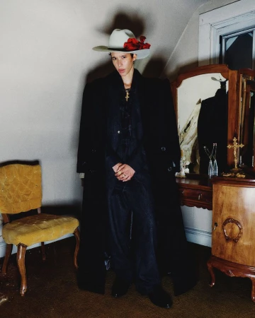 A person wearing a cowboy hat and long coat stands in an indoor setting next to an antique dresser and yellow upholstered chair.
