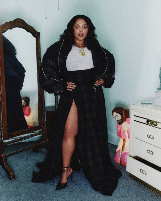 Adult female poses indoors with a confident expression, wearing a black bomber jacket, high-slit skirt, and high-heeled shoes.