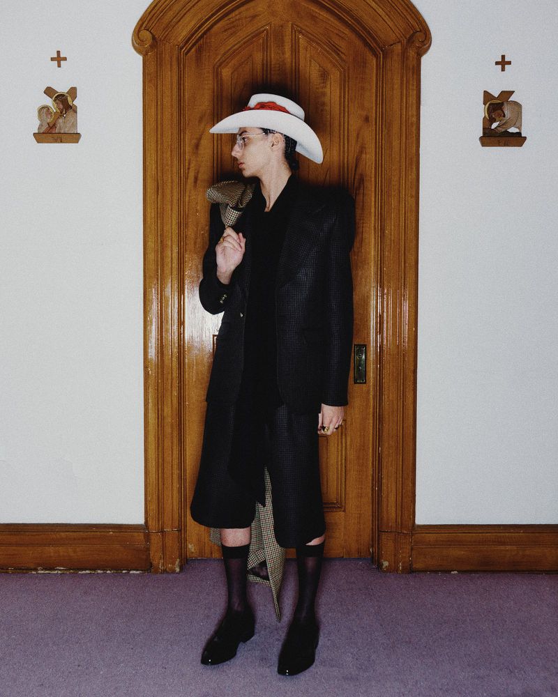 A stylish person in a dark suit and white cowboy hat poses in front of a wooden door, accented by religious-themed wall decor.