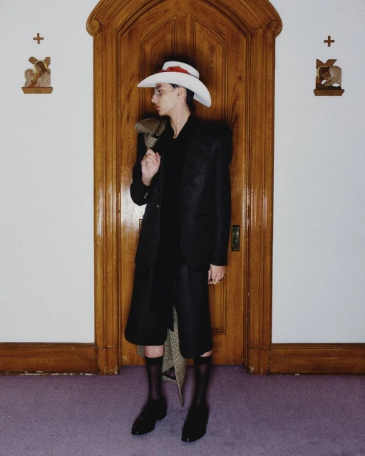 A stylish person in a dark suit and white cowboy hat poses in front of a wooden door, accented by religious-themed wall decor.