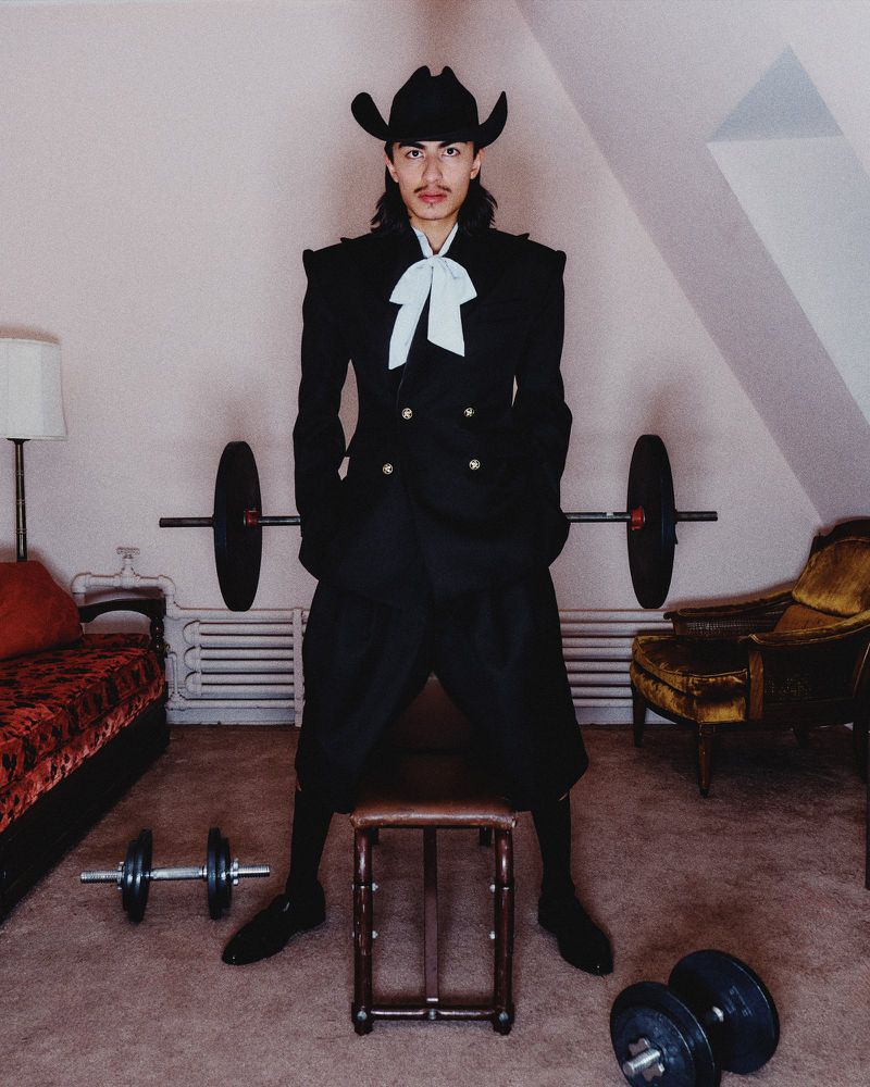 Person in black suit and cowboy hat stands next to workout equipment in indoor room with patterned carpet and painted walls.