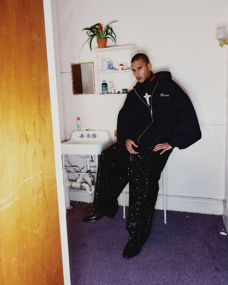 Stylish man poses in a black jacket and sequined pants in a white-walled room with a vintage sink and decor, creating a high-fashion image.