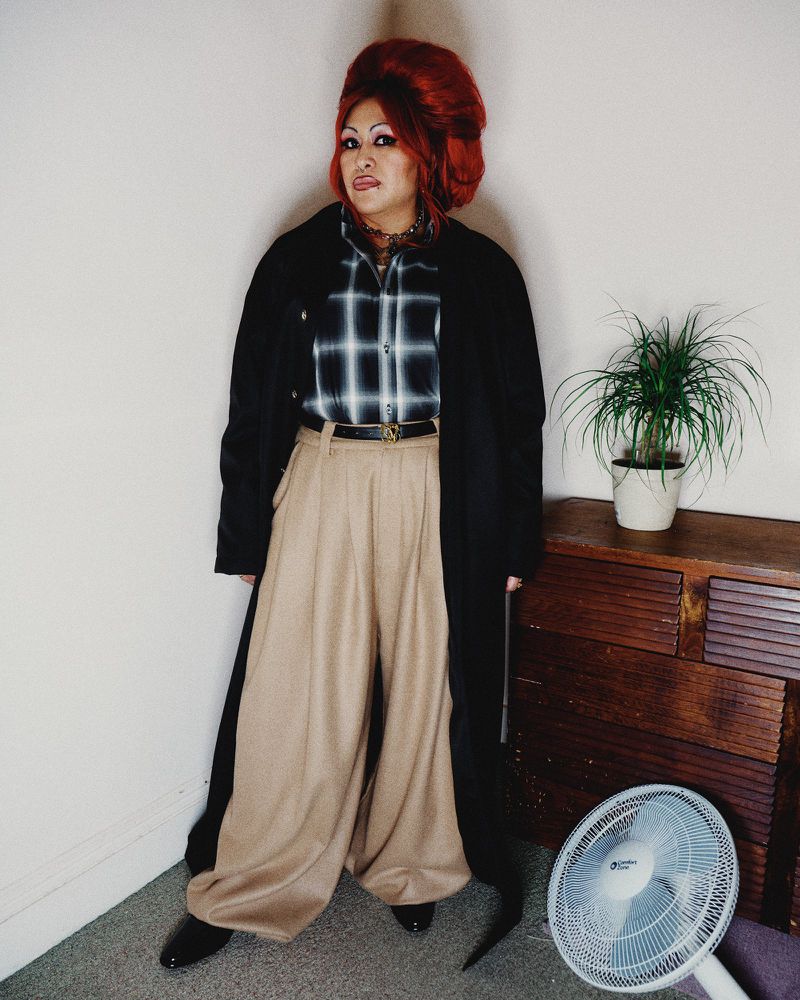 Full-length shot of an individual with high red hair wearing a plaid shirt, wide tan pants, and a black coat, indoors, looking at the camera.