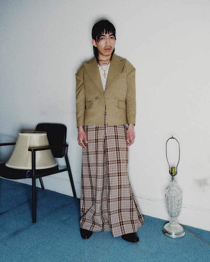 An Asian adult male stands in a photo studio wearing a blazer, plaid pants, rings, and a gold cross necklace against a white wall.