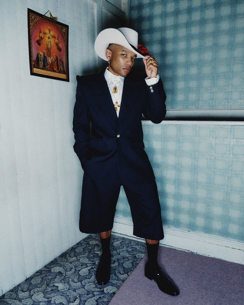 Stylish man in suit shorts, cowboy hat, and polished shoes poses in a corner. A framed painting hangs on the light blue wall.