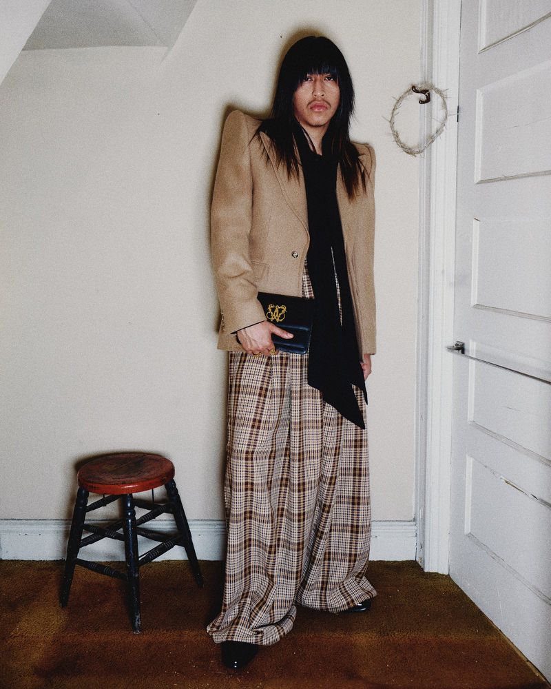 Adult posing indoors in a tan blazer, black top, and plaid pants, holding a black purse with a gold emblem.
