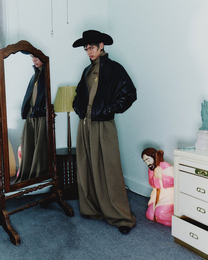 A person in oversized trousers, black jacket, and hat stands in a room next to a mirror, showcasing a fashion-forward and casual style.