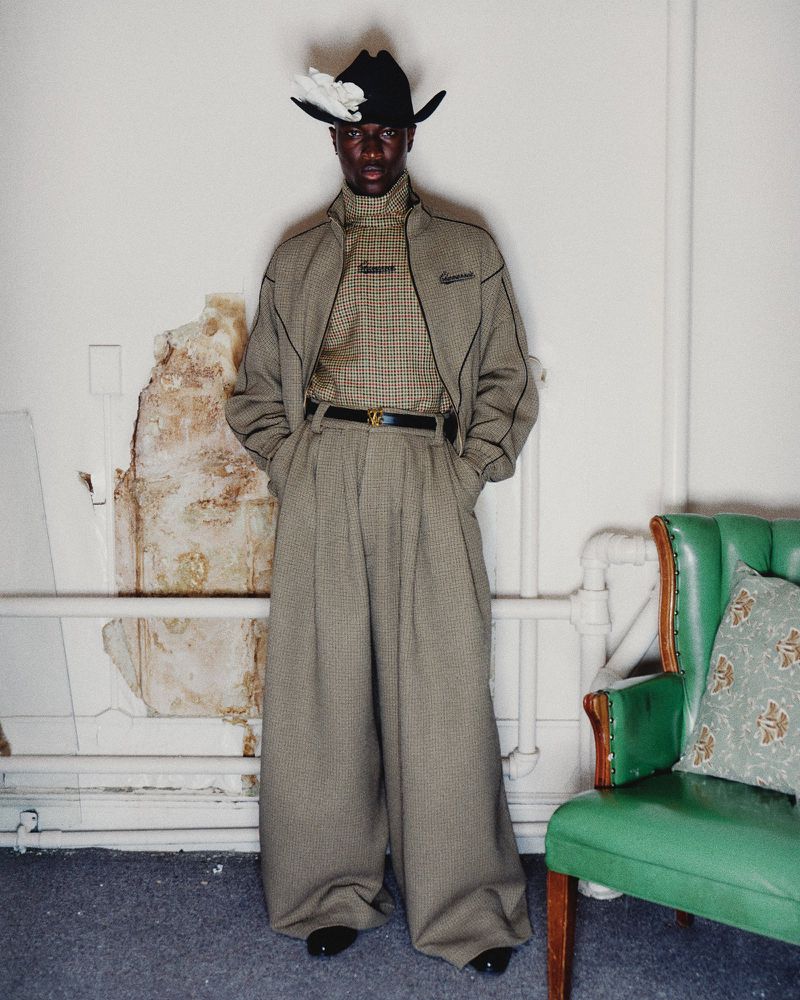 Stylish black man in cowboy hat, checked shirt, blazer, and wide-leg pants poses against a wall indoors. Vintage armchair to the side.