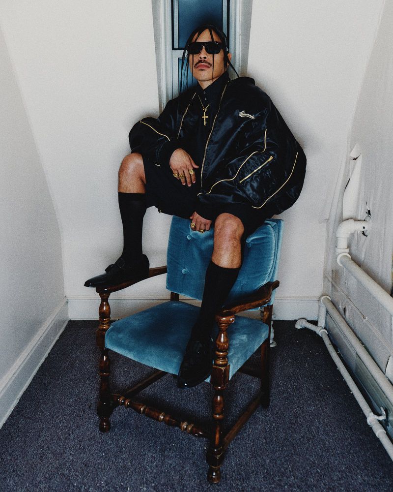 Stylish man in sunglasses and a jacket sits on a blue chair indoors, posing for a fashion shot with a mustache and cross necklace.