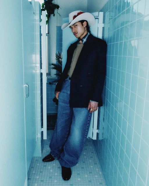 Young man in a cowboy hat and blazer poses in a tiled room, exuding a cool, casual vibe with a touch of retro fashion.