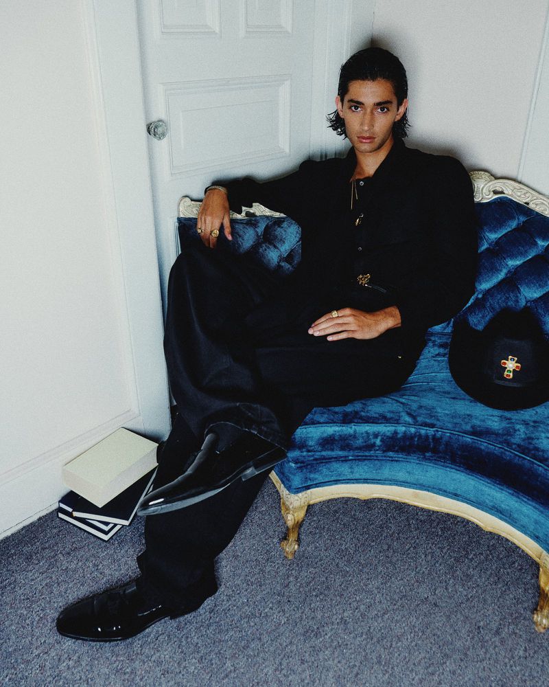 Portrait of a stylish adult male sitting on a tufted blue settee indoors in an all-black ensemble, with glossy dress shoes.