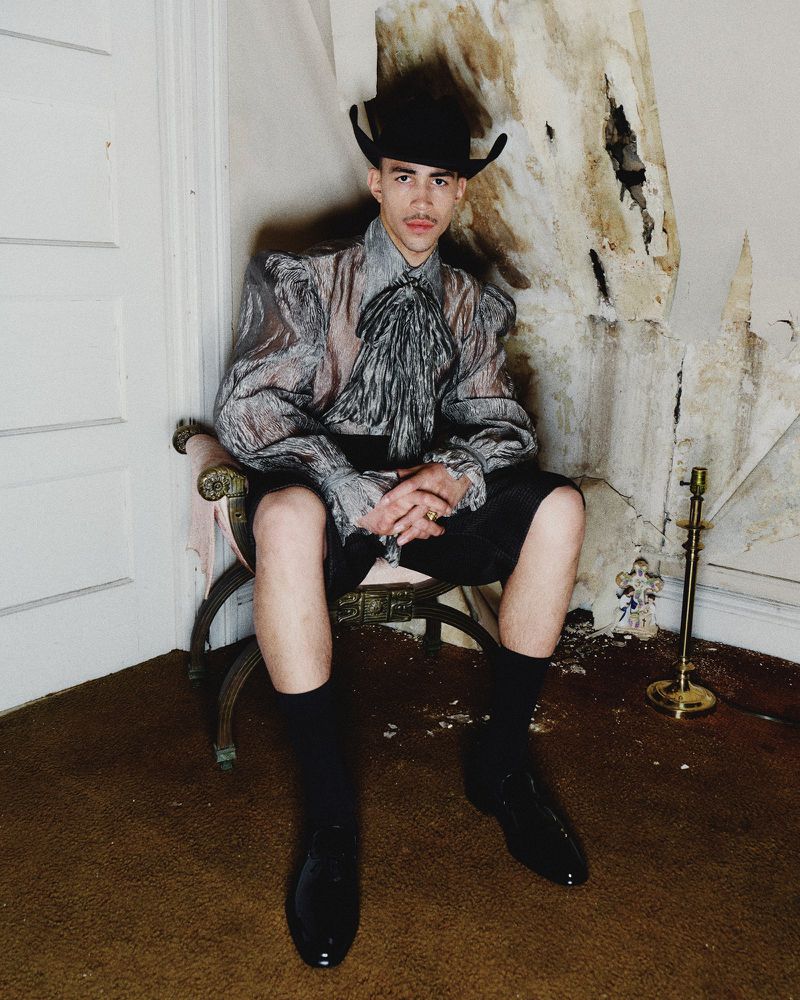A person with a mustache and a cowboy hat sits in a chair, wearing a see-through blouse and shorts, in front of a decaying wall.