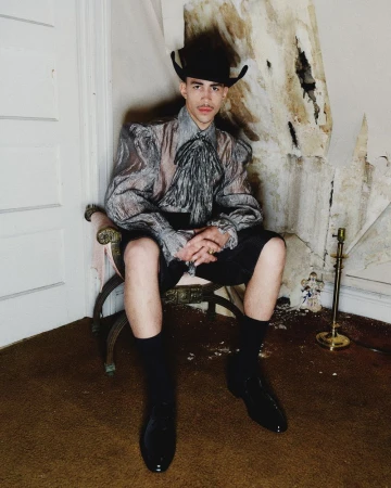 A person with a mustache and a cowboy hat sits in a chair, wearing a see-through blouse and shorts, in front of a decaying wall.
