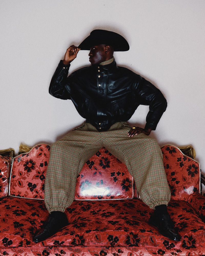 A Black adult male posing in a leather jacket, cowboy hat, and patterned pants, seated on a red floral sofa against a plain background.