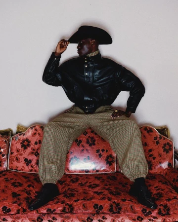 A Black adult male posing in a leather jacket, cowboy hat, and patterned pants, seated on a red floral sofa against a plain background.