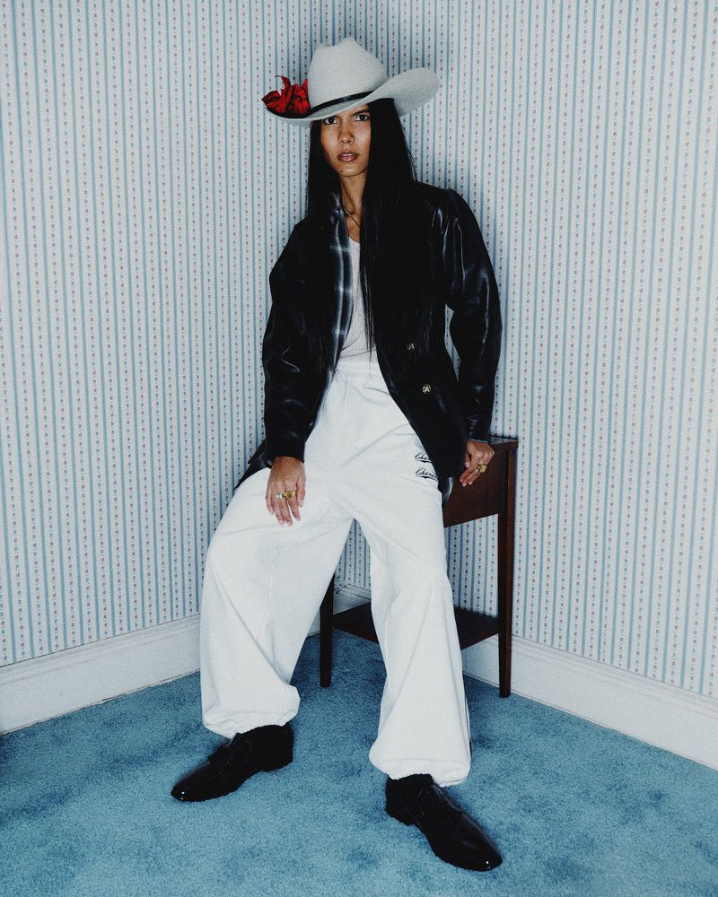 Full-shot of an Asian woman in a cowboy hat, black jacket, and white pants, posing in an indoor corner with blue-striped wallpaper.