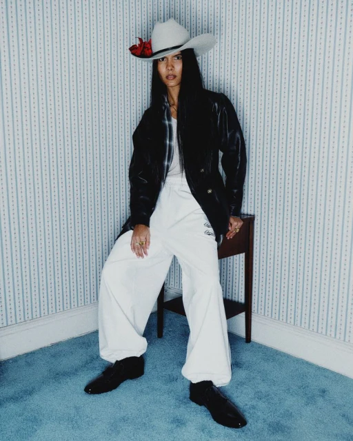 Full-shot of an Asian woman in a cowboy hat, black jacket, and white pants, posing in an indoor corner with blue-striped wallpaper.