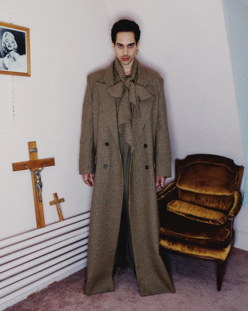 Adult male in a patterned overcoat with a large bow, standing in an interior with religious items and a brown velvet chair.