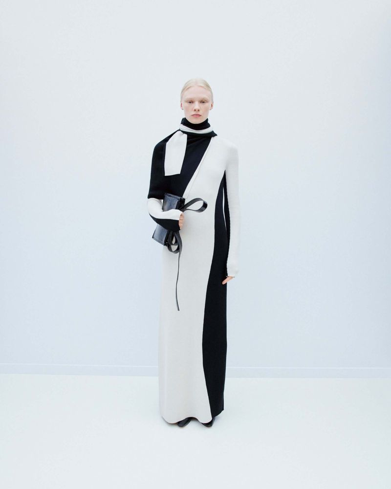 Elegant woman in a black and white long dress holding a black clutch, standing in a bright white studio setting, fashion forward.
