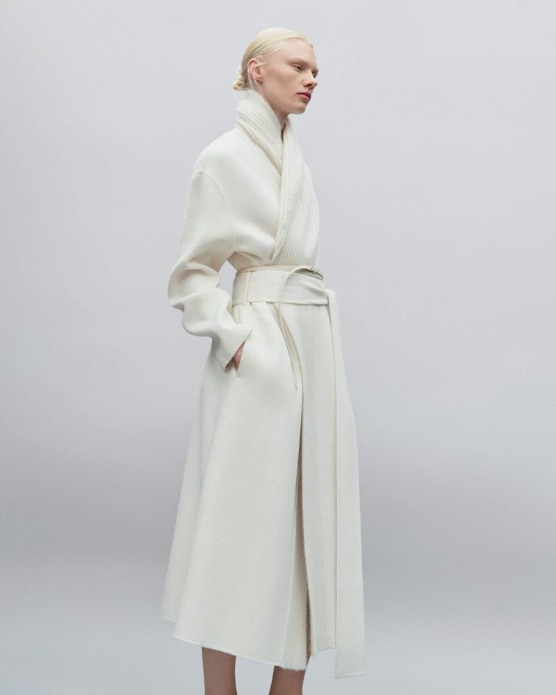 Elegant albino woman in full-length white coat with matching belt and minimalist styling, posing against a pale grey studio backdrop.