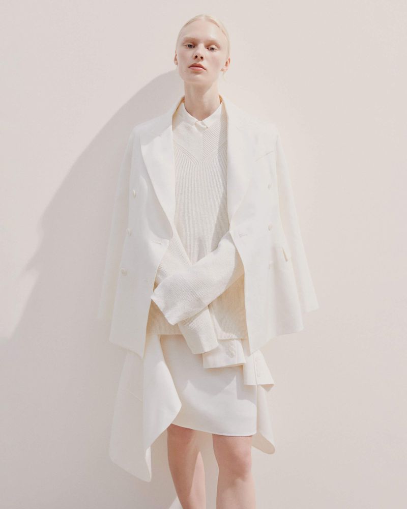Fashionable woman in white blazer and skirt poses against a cream wall, casting a soft shadow, in a minimalist editorial style.