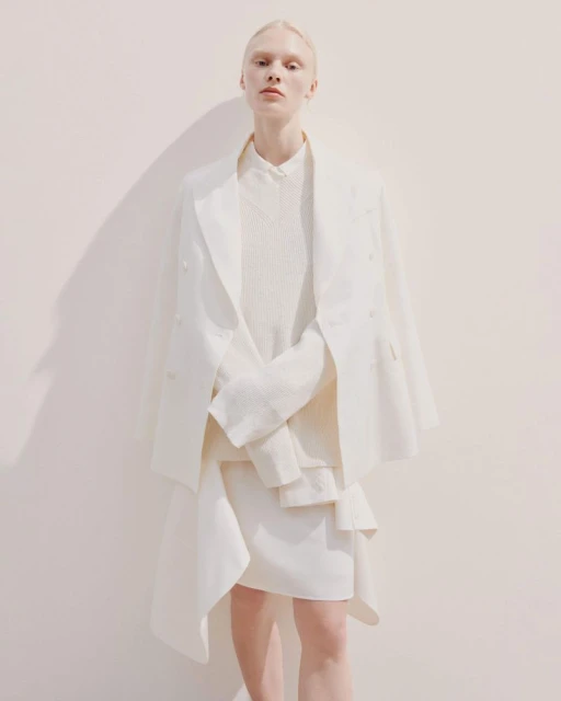 Fashionable woman in white blazer and skirt poses against a cream wall, casting a soft shadow, in a minimalist editorial style.